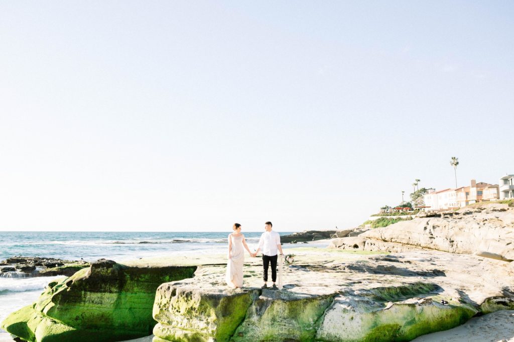 A married couple standing on a cliff edge in La Jolla, San Diego, holding hands and smiling at each other