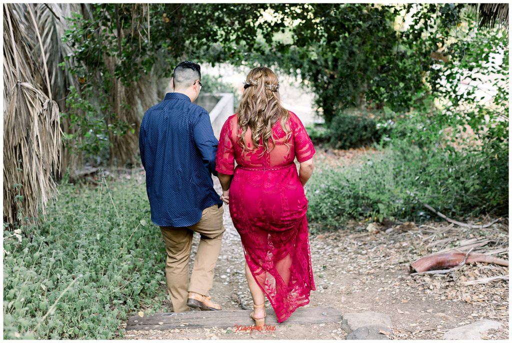 An image of an interracial couple (an Asian male wearing a dark blue button down shirt and khaki pants and a Latina female wearing a long red dress) holding hands and walking away from the camera