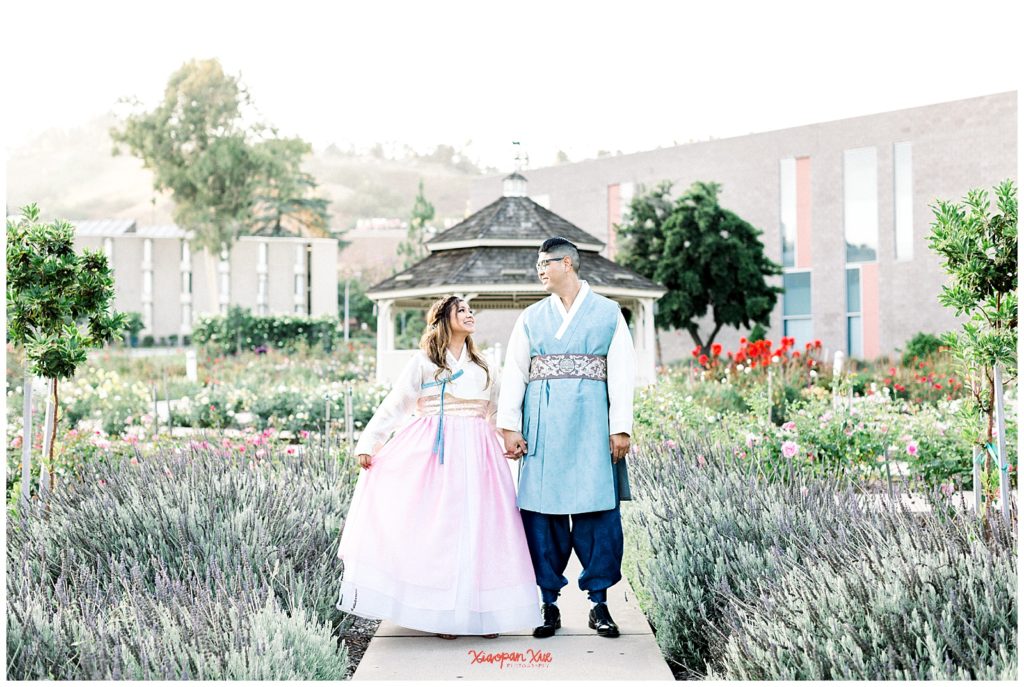 An image of an interracial couple (an Asian male and a Latina female) in traditional Korean hanbok wear (pink and blue being the main colors) holding hands and smiling at each other; they are in a rose garden