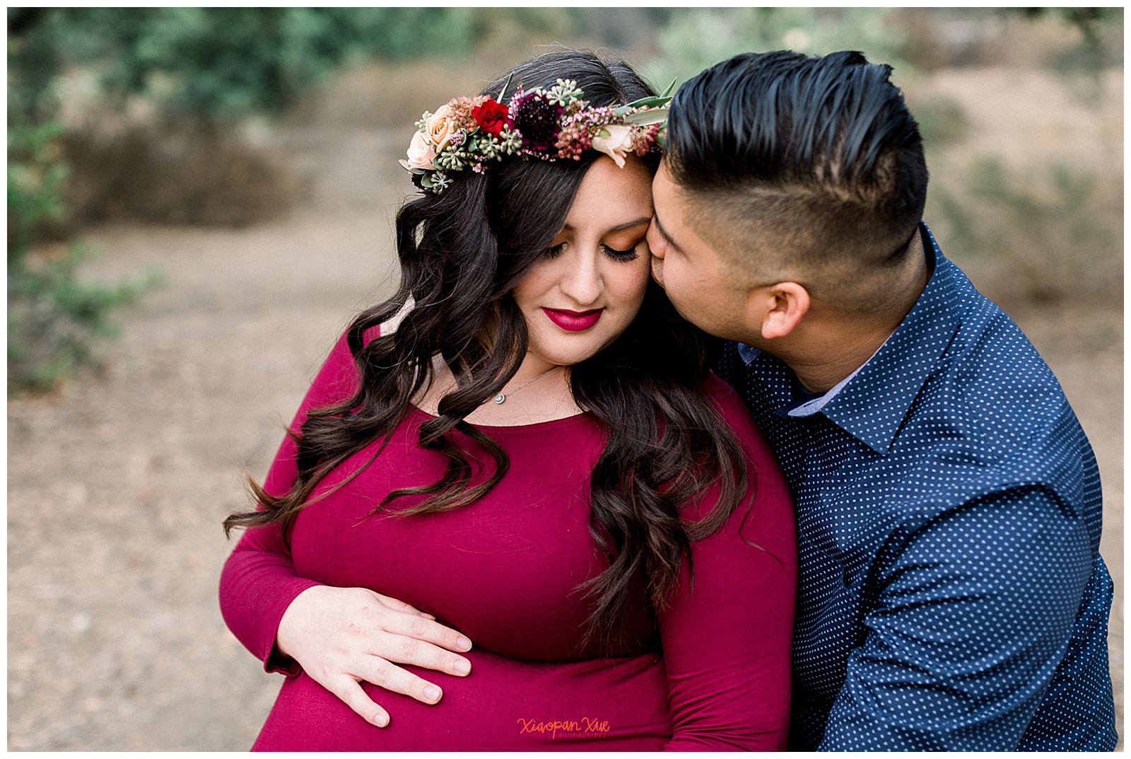 An image of an interracial couple (an Asian male and a Latina female). The male is hugging her from the side and kissing her temple. The female has her hand on her expectant belly.